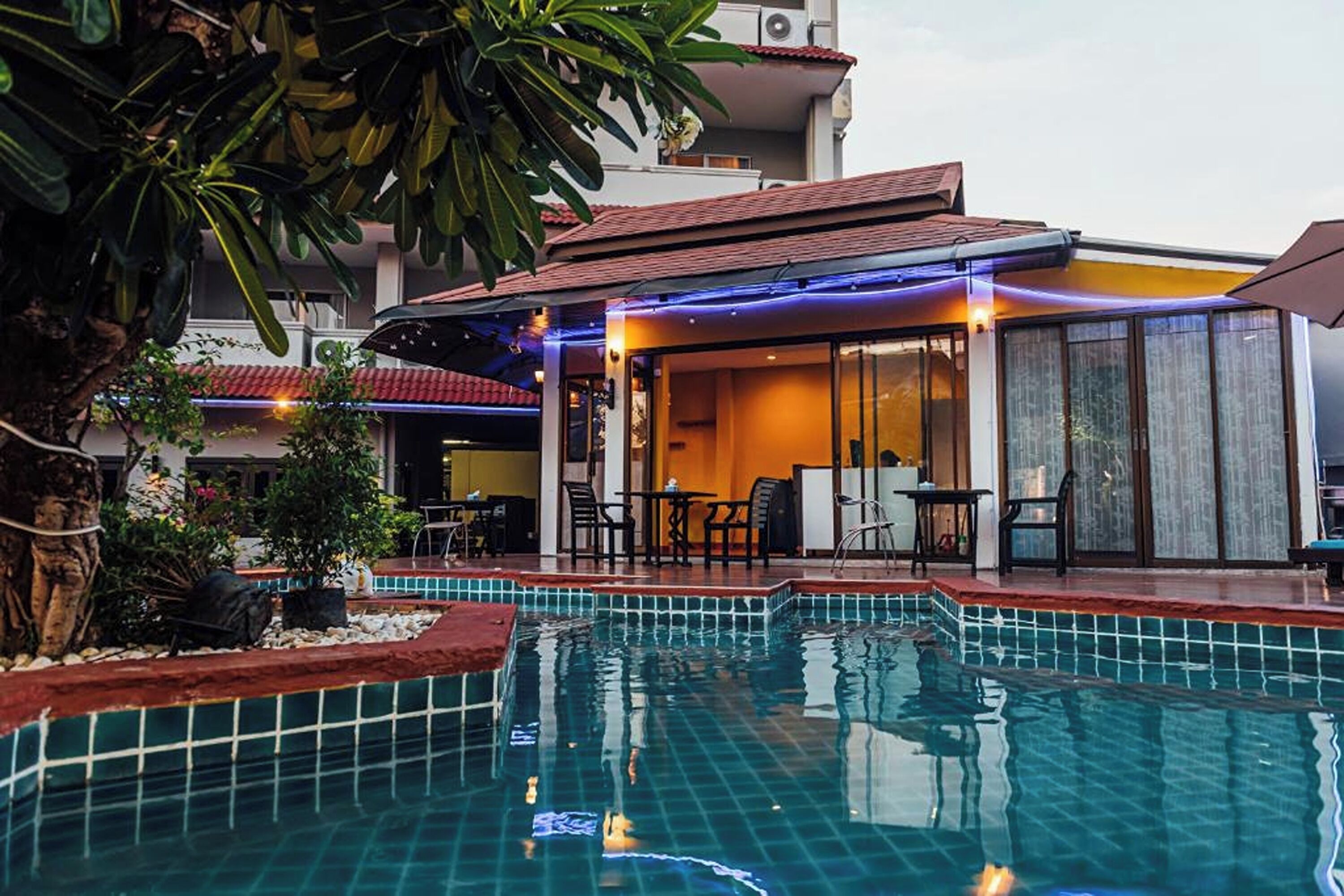 A pool area with a tiled edge, a covered seating area with chairs and tables, and a building with a red roof in the background.