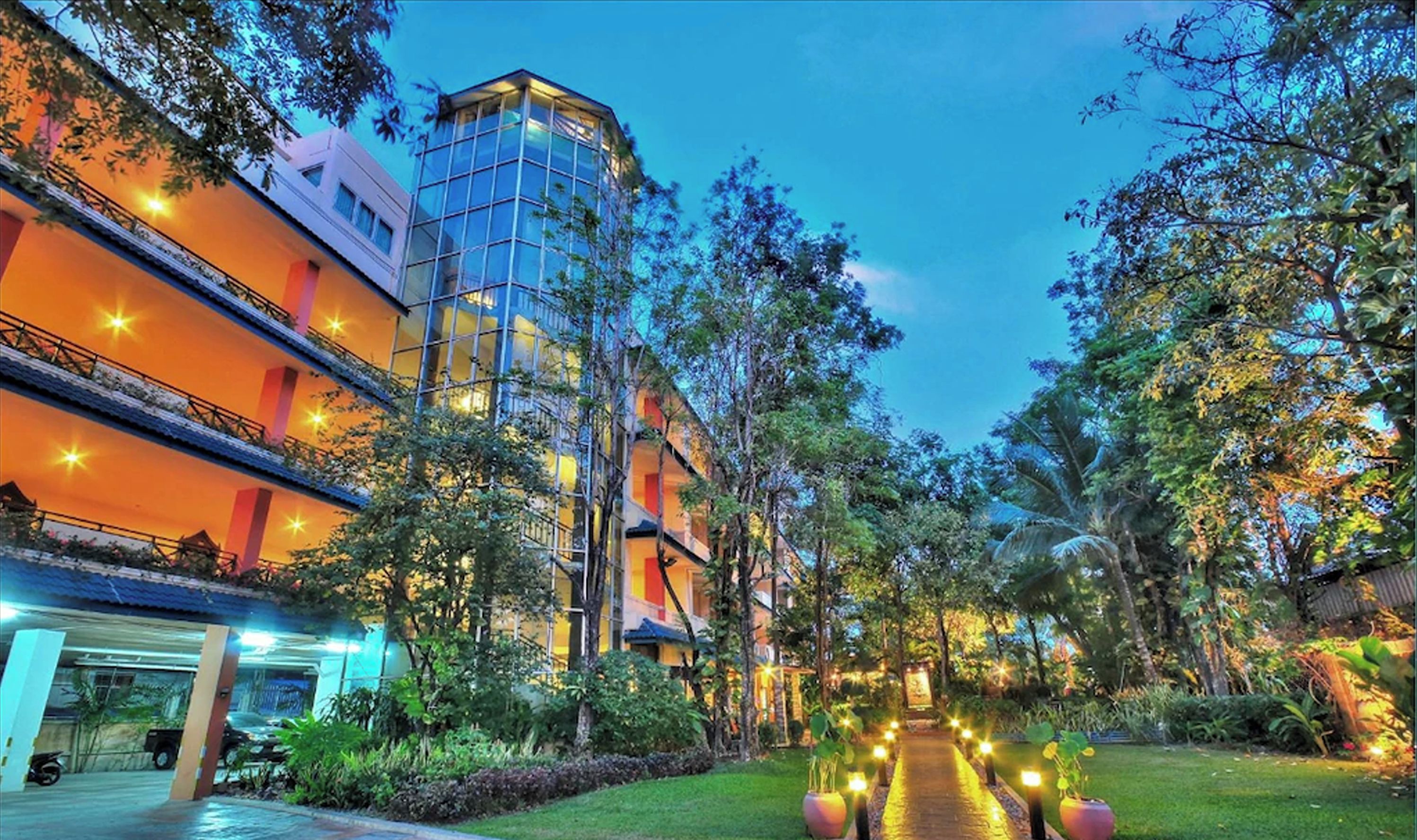 A multi-story hotel with a glass facade, surrounded by lush greenery and illuminated pathways.