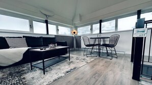 Ein modernes Zimmer mit einer Couch, einem Couchtisch und Sesseln. Große Fenster und eine Heizung sind vorhanden.