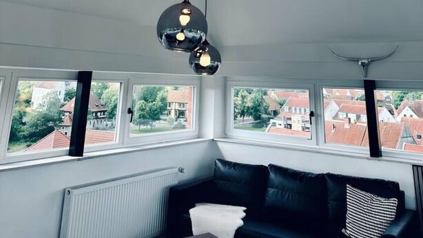 Ein modernes Wohnzimmer mit einem schwarzen Ledersofa, einem weißen Zierkissen und einem Kissen mit schwarz-weißem Muster. Drei Fenster bieten Blick auf die Nachbarschaft.