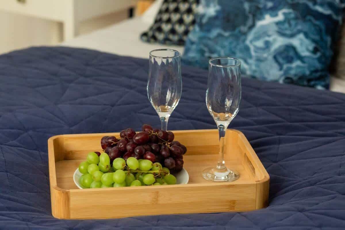 A wooden tray with a selection of grapes and two empty wine glasses on a bed with a blue quilted cover.