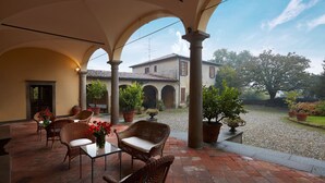 A covered patio with wicker furniture, potted plants, and a view of a building with arches.