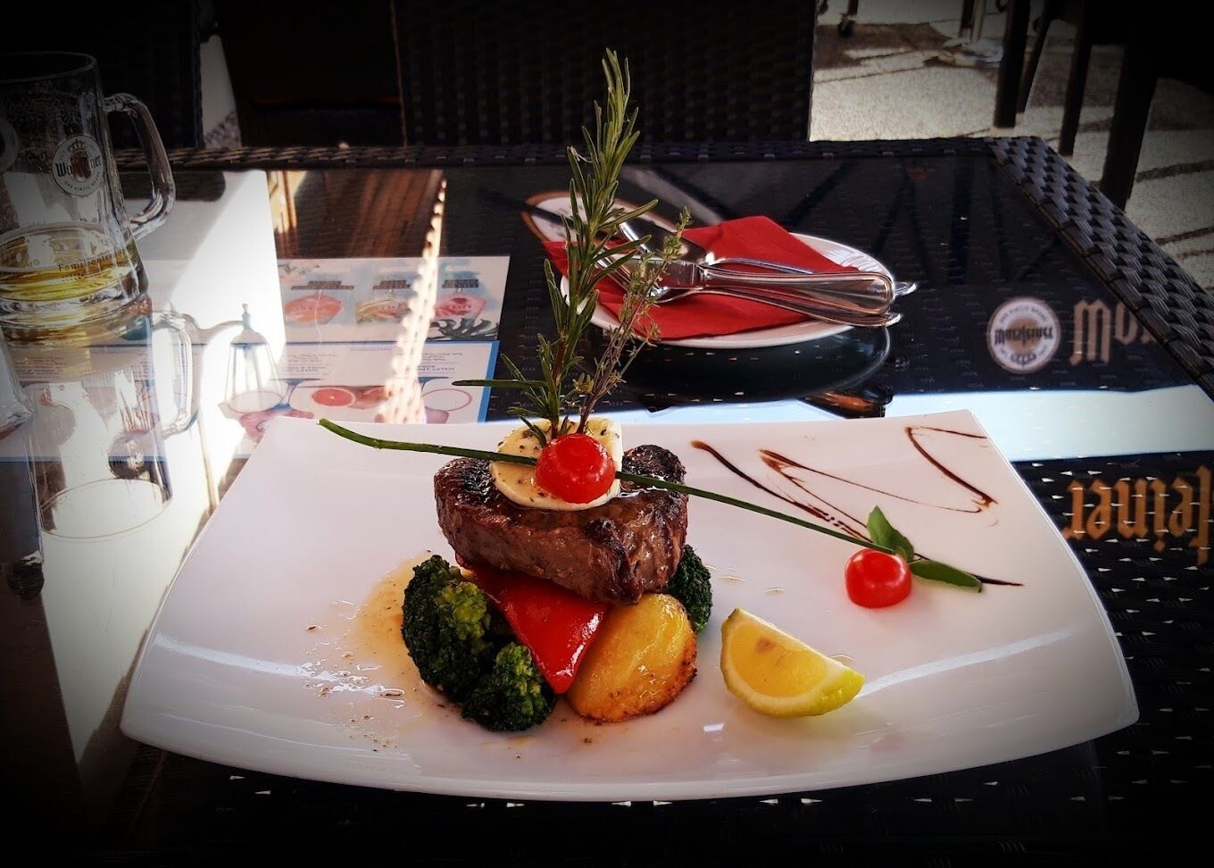 A plated dish with steak, broccoli, and cherry tomatoes, garnished with herbs, on a table with a menu and a glass of beer.