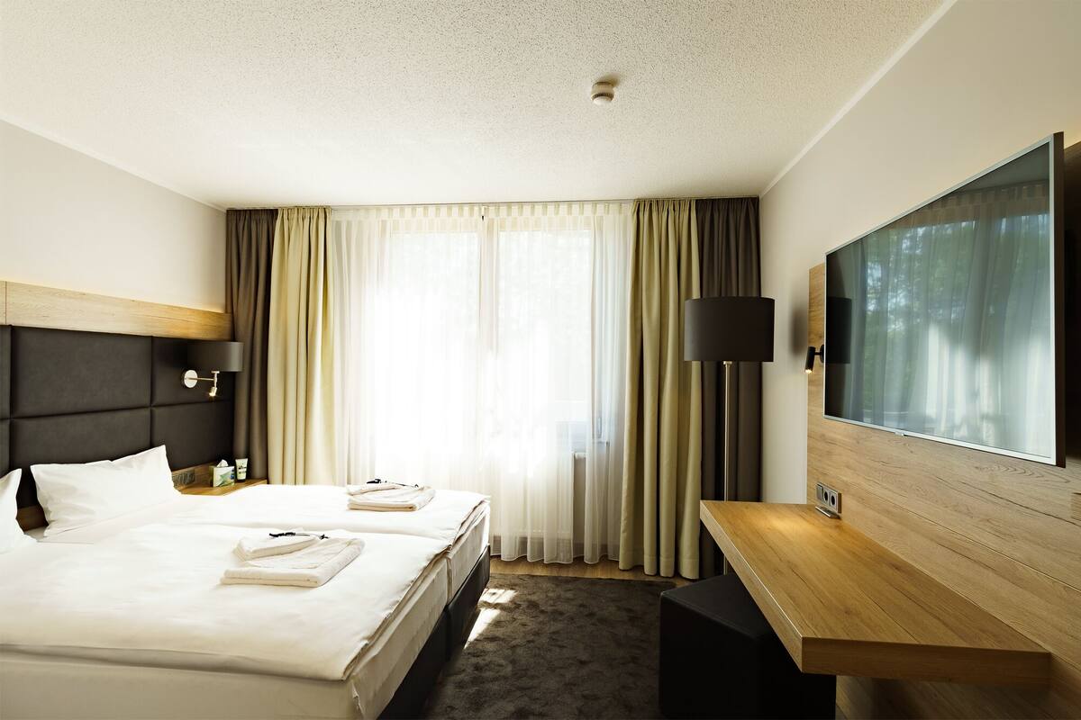 Habitación de hotel con cama, dos mesitas de noche, un escritorio, televisor de pantalla plana y un gran ventanal con cortinas.