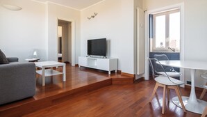 Uma sala de estar moderna com uma TV de tela plana, um sofá cinza, uma mesa de centro branca e uma área de jantar com uma mesa e cadeiras brancas.