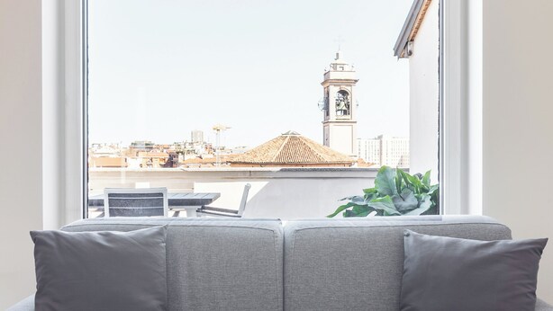 Um sofá cinzento com almofadas, uma janela com vista para um horizonte urbano e uma planta no parapeito.
