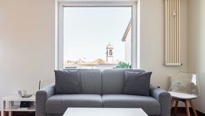 Uma sala de estar moderna com um sofá cinzento, uma mesa de centro branca e vista para o horizonte urbano através de uma janela grande.