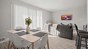 A modern living room with a dining area, a grey sofa, and a TV mounted on the wall. - Stylish & Modern Unit  Prime Location & Comfort (Rochester Hills)