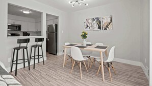 A modern kitchen with white cabinets, a stainless steel refrigerator, and a microwave. There is a dining area with a wooden table and white chairs. - Stylish & Modern Unit  Prime Location & Comfort (Rochester Hills)