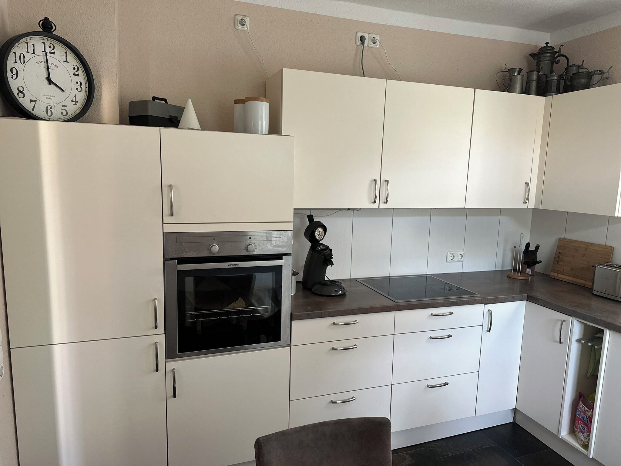 A modern kitchen with white cabinets, a built-in oven, a coffee machine, and a clock on the wall.