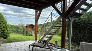 Une terrasse couverte avec une table en verre et un fauteuil en osier, donnant sur une maison de jeu en bois et un escalier menant à une terrasse.