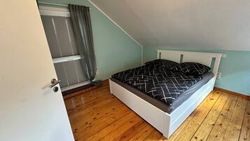 A bedroom with a sloped ceiling, a bed with a patterned bedspread, wooden flooring, and a window with curtains.