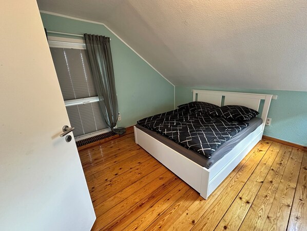 A bedroom with a sloped ceiling, a bed with a patterned bedspread, wooden flooring, and a window with curtains.