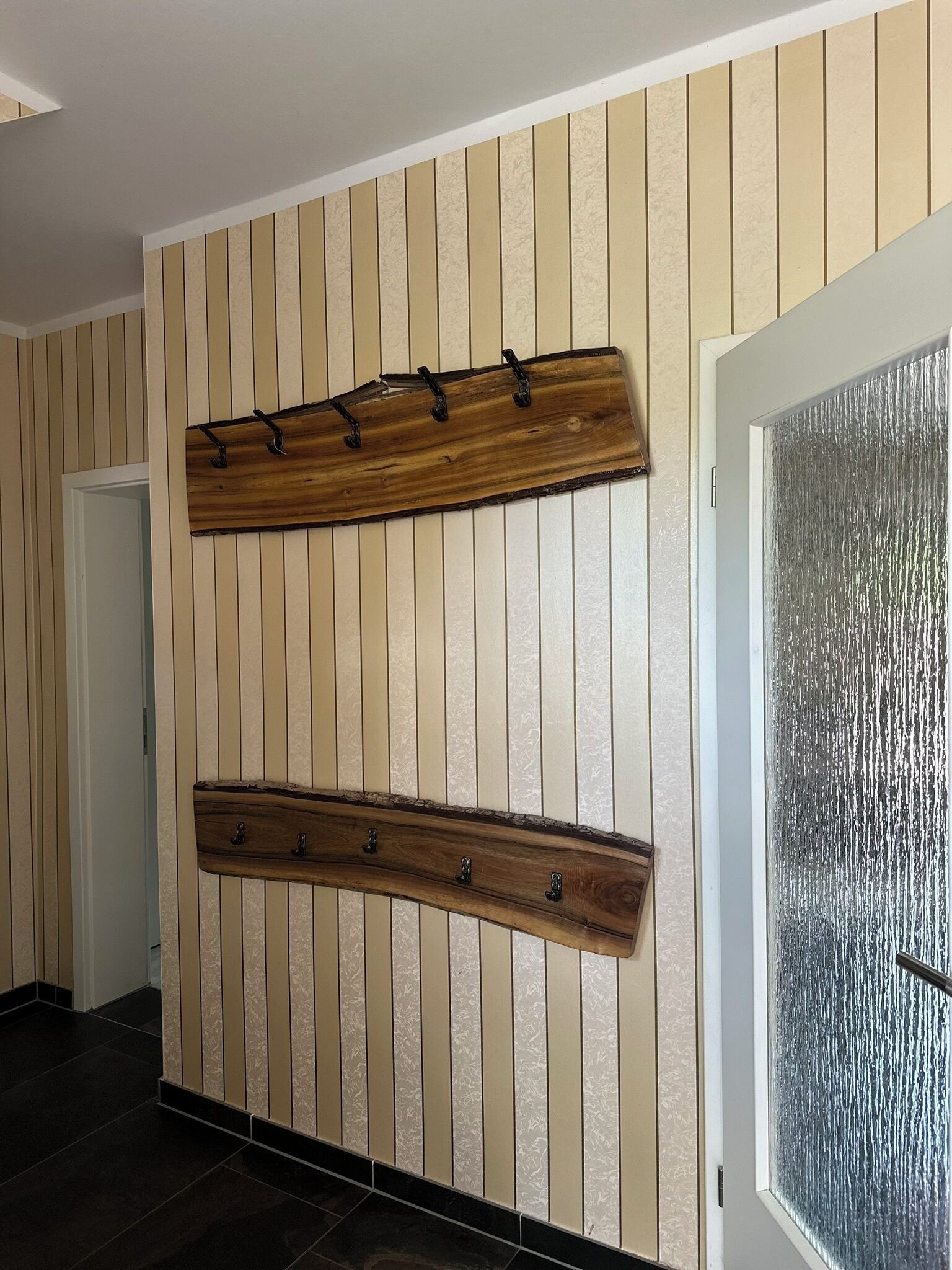 Un pasillo con papel tapiz de rayas verticales, un perchero de madera fijado a la pared y una puerta con panel de vidrio texturizado.