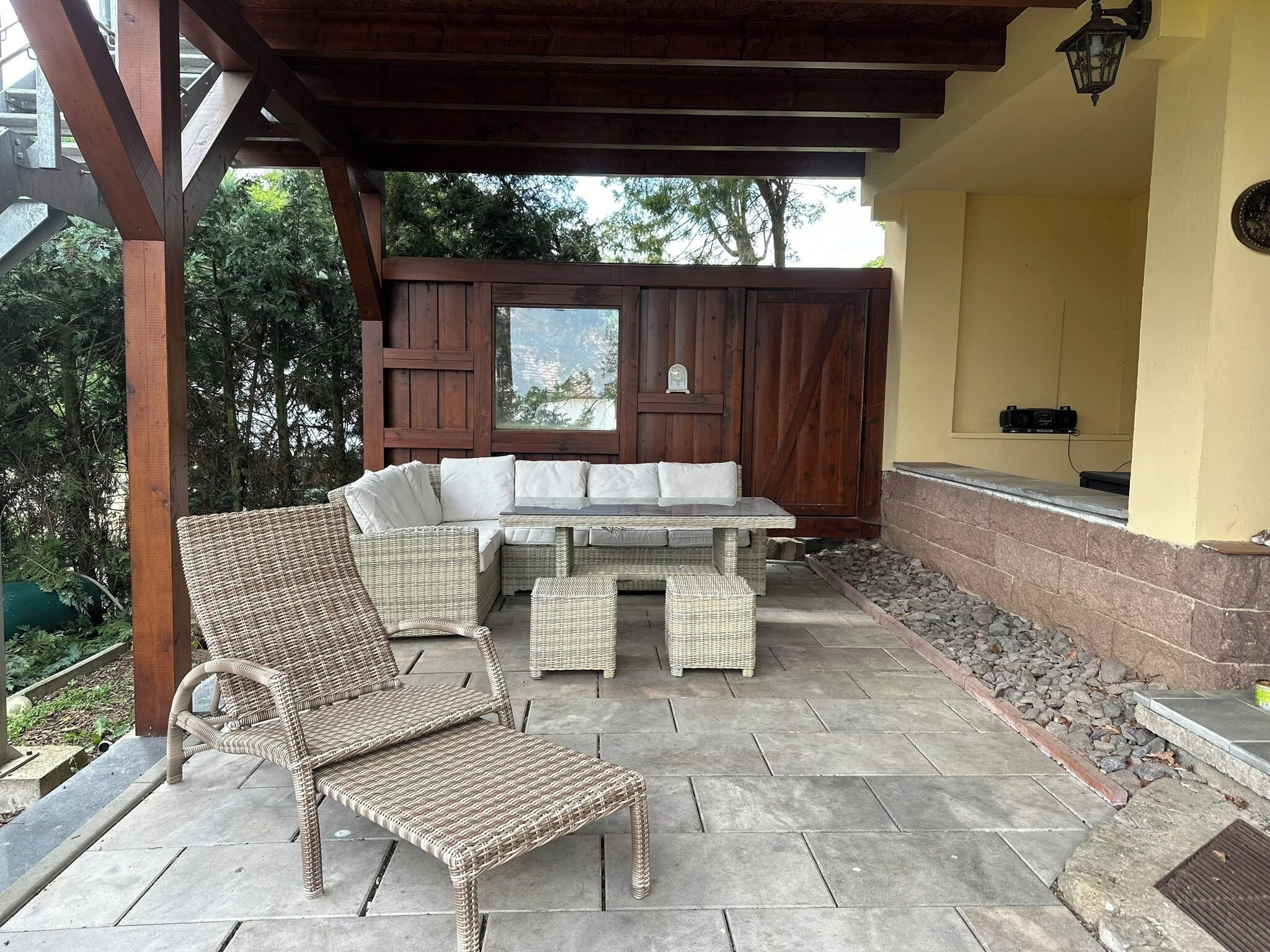 Un patio al aire libre con mobiliario de mimbre, incluyendo un sofá y sillas, bajo una pérgola de madera.
