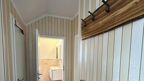 Un couloir aux murs rayés, une poutre en bois avec des crochets, et une porte donnant sur une salle de bain.