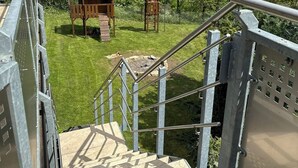 Un escalier mène à une zone herbeuse où se trouvent une structure de jeu en bois et un petit abri.