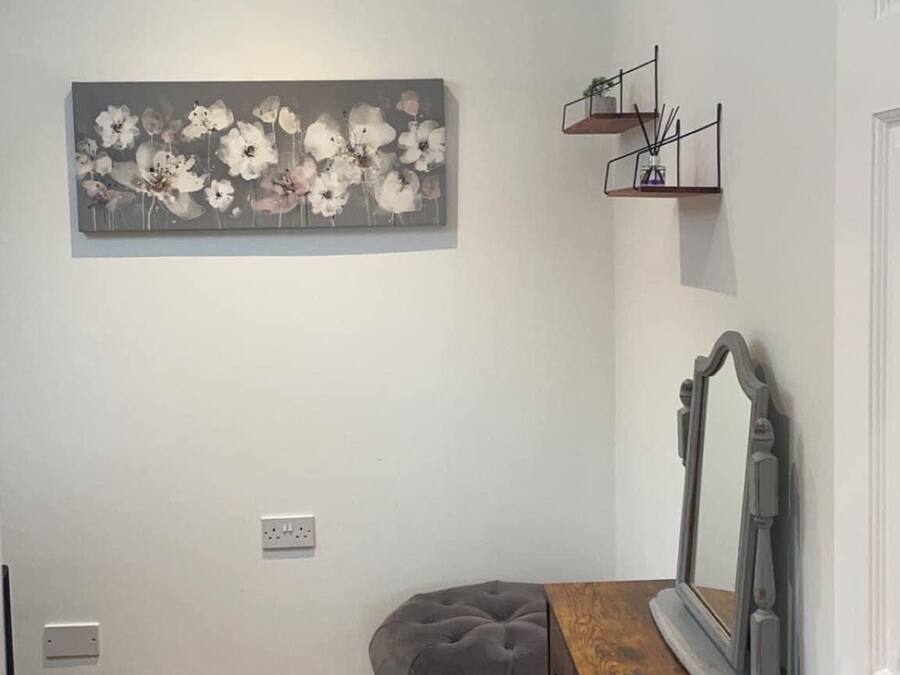 Una habitación con un otomano capitonado gris, un aparador de madera con espejo, un cuadro floral gris en la pared y una pequeña estantería con plantas.