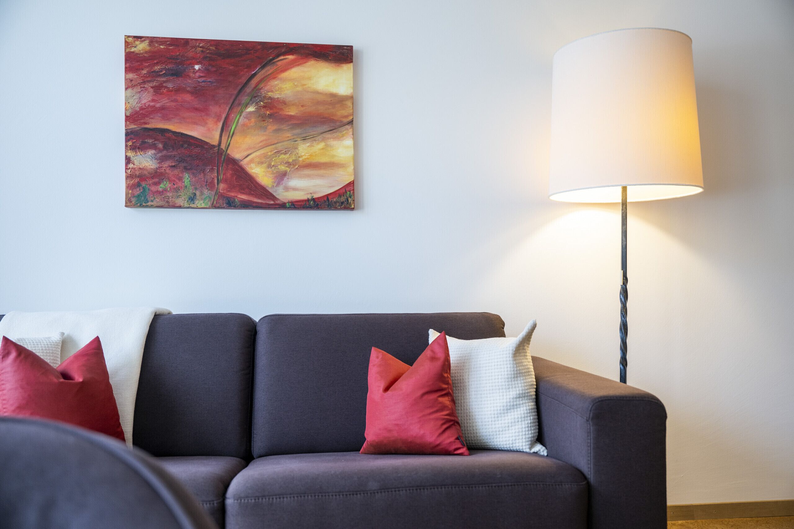 Una sala de estar moderna con un sofá gris oscuro, cojines rojos y blancos, un cuadro abstracto grande y una lámpara de pie.
