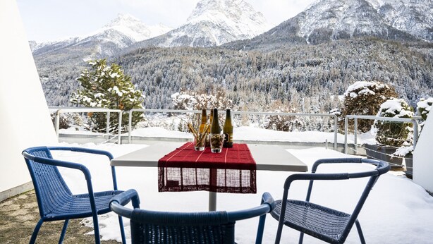 A balcony with a table set for two, overlooking snow-covered mountains. - CHASA TULAI F83: BEAUTIFULLY RENOVATED STUDIO IN HOLIDAY PARK TULAI F83 (Scuol)