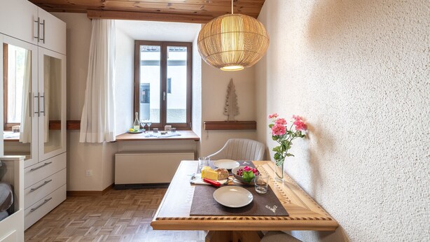 A cozy kitchen with wooden cabinets, a dining table set with food, a hanging light fixture, and a window with curtains. - Chasa Vi Studio: located in the center of Scuol (Scuol)