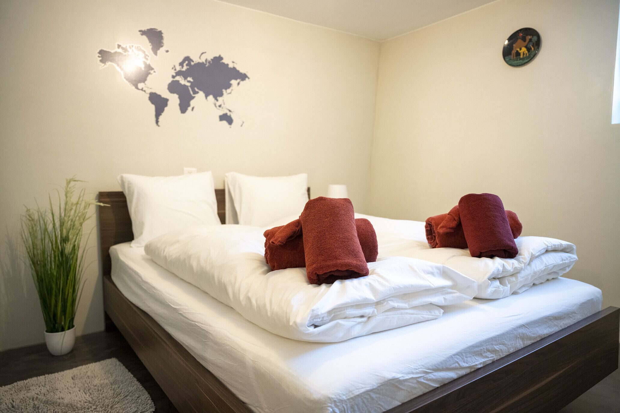 Una cama bien tendida con sábanas blancas y dos toallas rojas, un mapa mundial en la pared y una planta pequeña al lado de la cama.