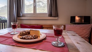 Une table à manger avec une assiette de nourriture et des verres remplis d’une boisson rouge, un téléviseur et une vue sur les montagnes par la fenêtre.
