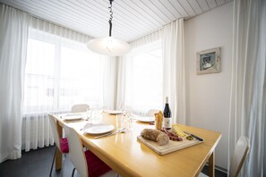 A dining area with a wooden table set for two, featuring a bottle of wine, bread, cheese, and cured meats.