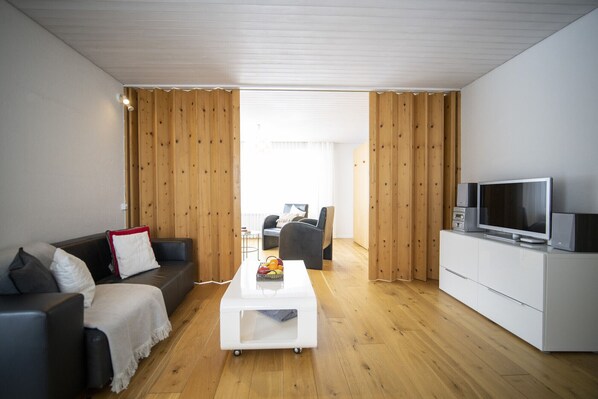 A modern living room with a wooden floor, a white coffee table, a grey sofa, a black armchair, and a TV on a white cabinet.