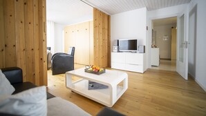 Ein modernes Wohnzimmer mit Holzboden, einem weißen Sofa, einem schwarzen Sessel, einem weißen Couchtisch mit Früchten, einem Fernseher auf einer weißen Kommode und einem Holzschrank.