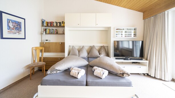 A modern bedroom with a bed, a bookshelf, a desk, and a TV. - Chasa Trü No. 8: Idyllic 2.5-room flat in the Trü holiday village (Scuol)