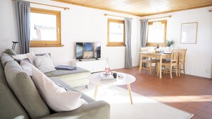 A living room with a sofa, TV, dining table, chairs, and a small coffee table. - Chasa Fabulusa: Modern 2.5-room apartment in the centre of Ftan (Ftan)
