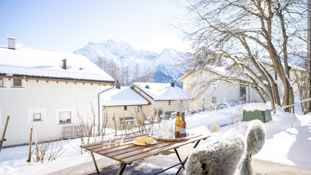 A wooden deck with a table set for a meal, overlooking a snowy landscape with houses and mountains in the background. - Chasa Fabulusa: Modern 2.5-room apartment in the centre of Ftan (Ftan)