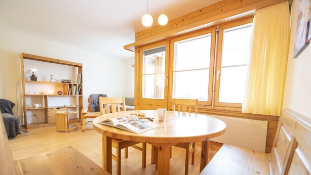 Une pièce chaleureuse avec une table à manger en bois, des chaises, une bibliothèque et de grandes fenêtres avec des rideaux.