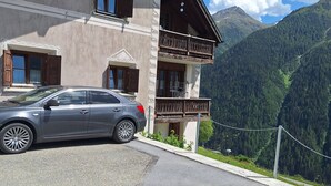 A two-story building with a car parked in front, a balcony, and a mountainous backdrop.