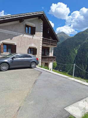 A two-story building with a car parked in front, a balcony, and a mountainous backdrop.