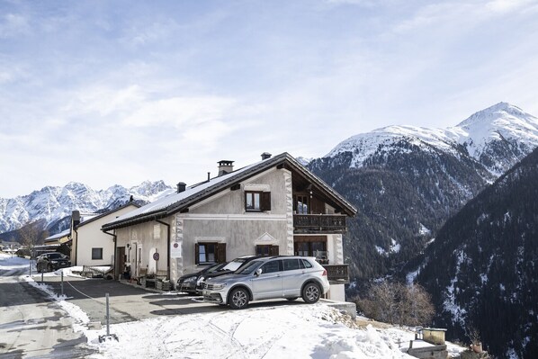 A two-story building with a balcony, parked cars, and snow-covered mountains in the background. - Chasa sur Chafarrer: Lovely 2.5 attic apartment in Guarda. (Guarda)