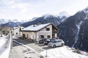 Un bâtiment à deux étages doté d’un balcon, avec des voitures garées et des montagnes enneigées en arrière-plan.