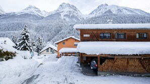 Ein Holzbau mit Garage und einem darin abstellendem Auto, umgeben von schneebedeckten Bäumen und Bergen im Hintergrund.