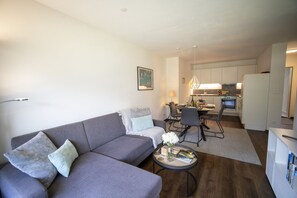 A modern living room with a grey sofa, a dining area with a table and chairs, and a kitchen with white cabinets and a built-in oven. - Brentschpark No. 33: Charming renovated 2.5-room flat in the quiet located Brentschpark (Scuol)