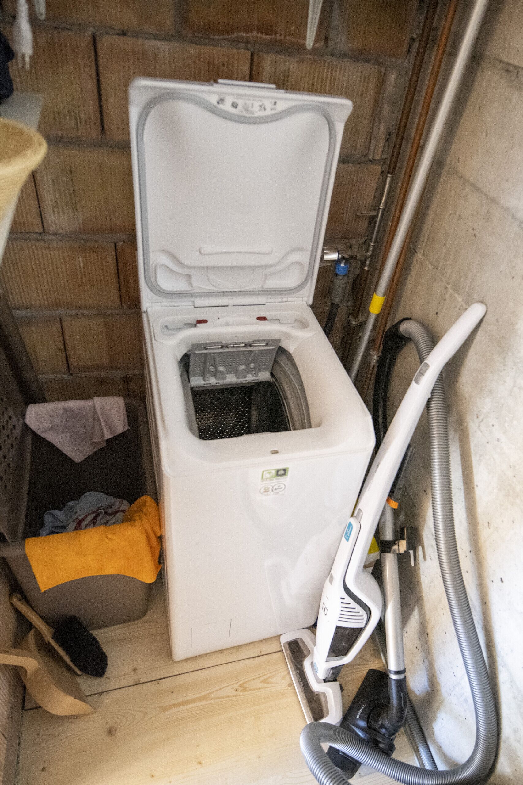 A compact, white, wall-mounted washing machine in a small, enclosed space with a wooden floor and a brick wall.
