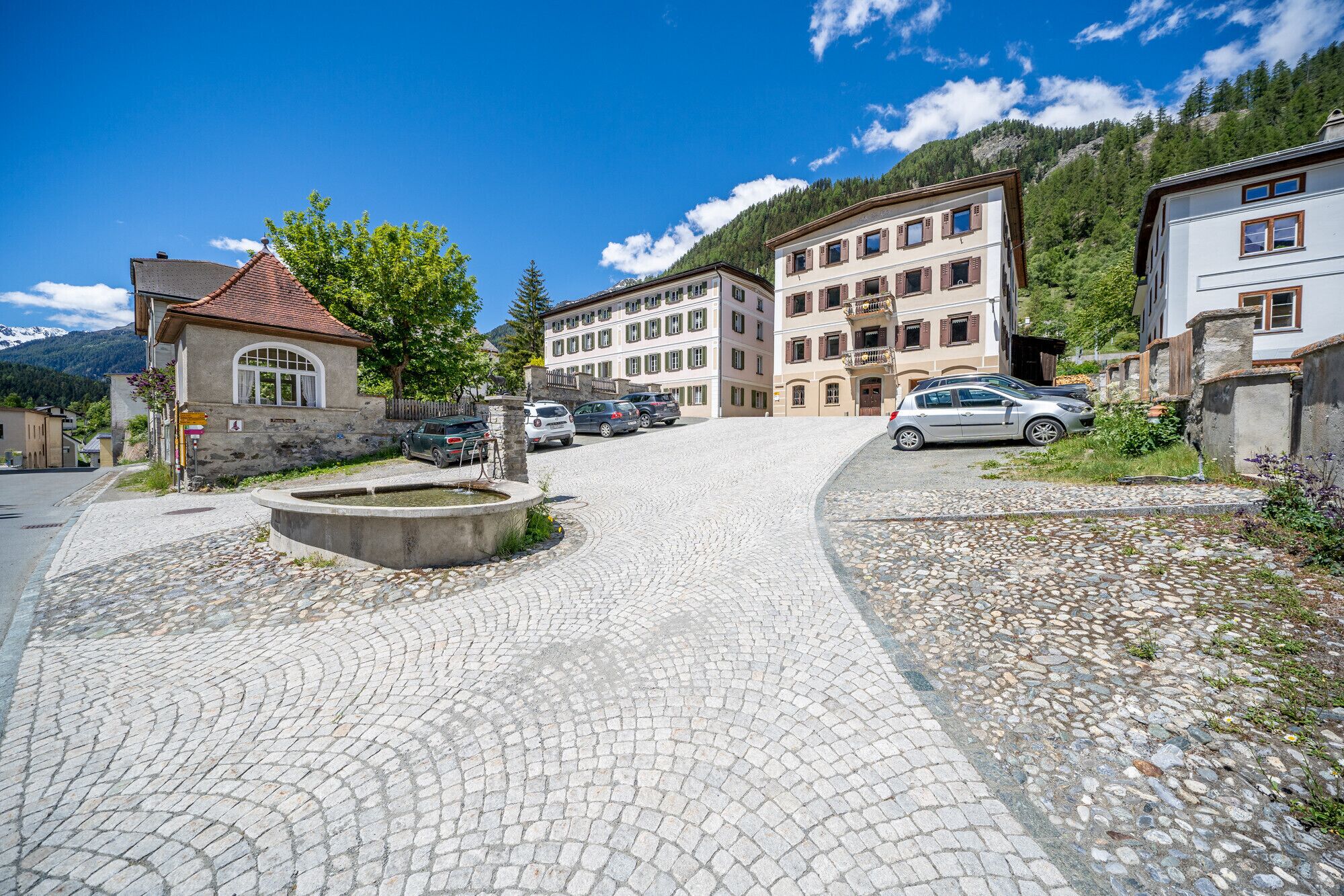 Calle adoquinada que lleva a un edificio con una fuente, coches aparcados y montañas al fondo.