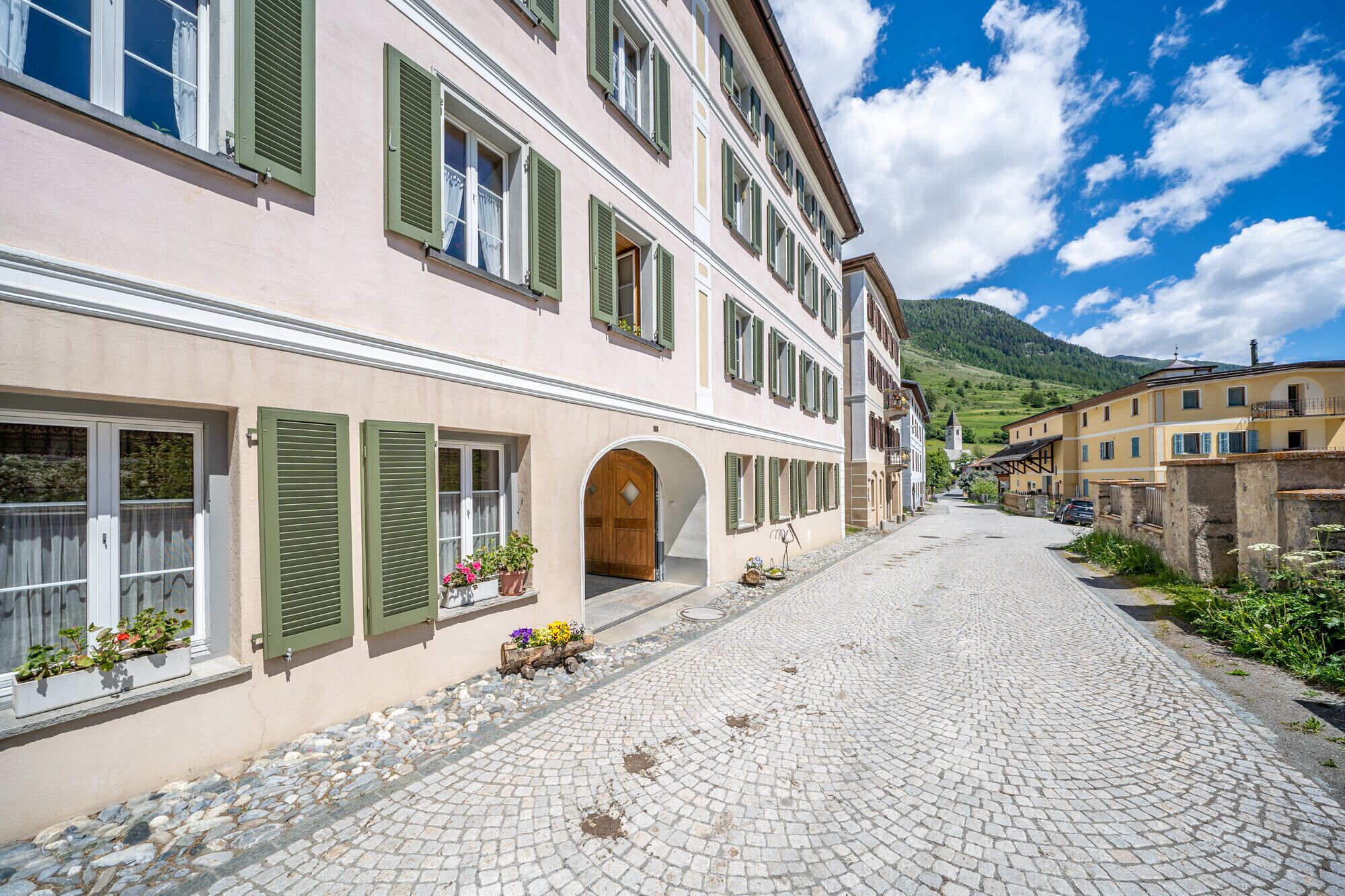 A cobblestone street lined with traditional buildings, featuring arched doorways and green shutters.