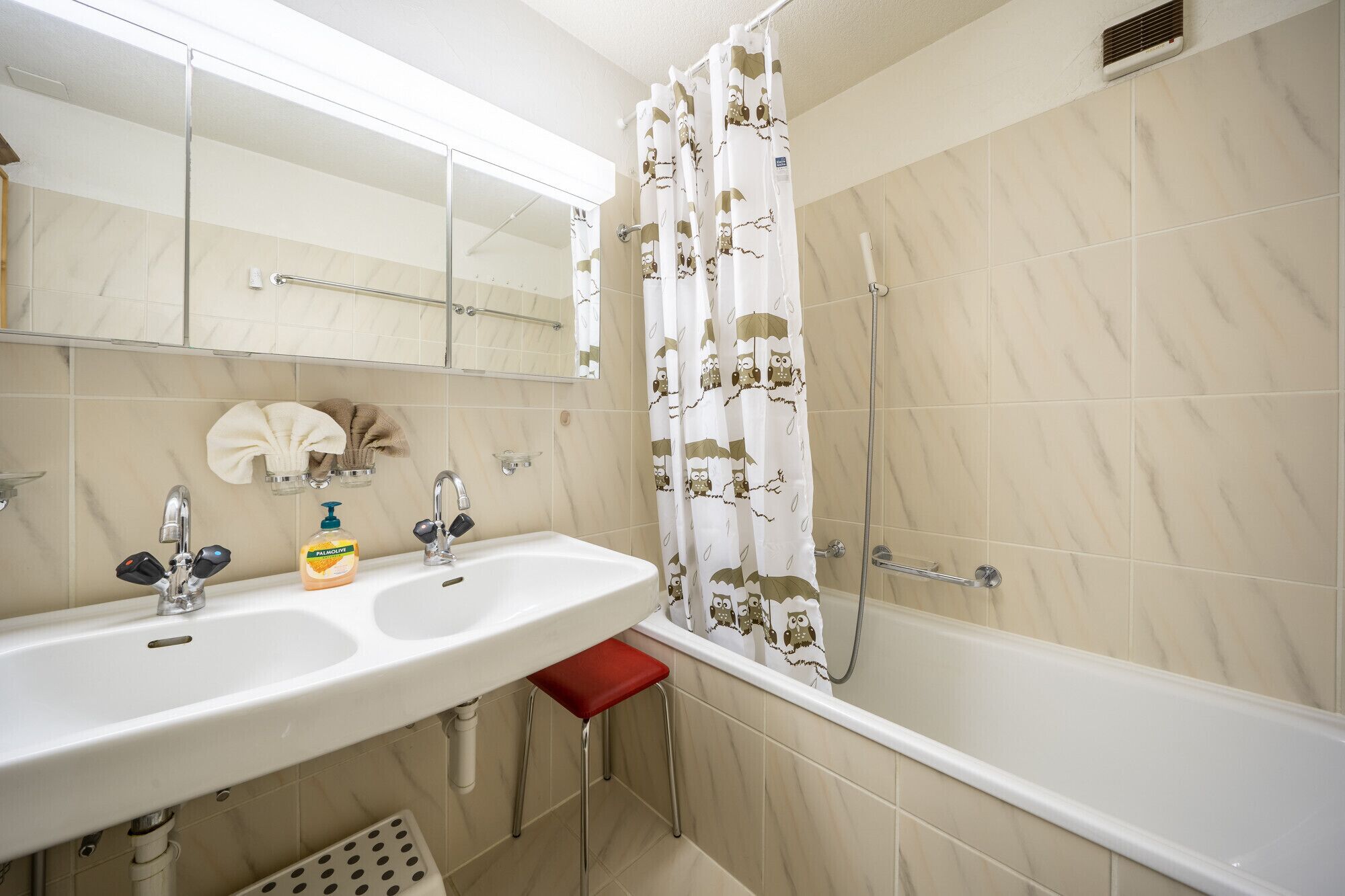 A bathroom with a white sink, two faucets, a mirror, a red stool, and a shower with a white curtain featuring owl patterns.