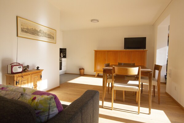 A room with a wooden dining table, chairs, a sofa, a wooden cabinet, a television on the wall, and a framed picture. - CHasa Avant Muglins: Garden flat with a view (Scuol)