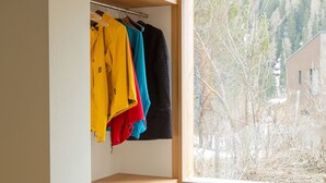 Un rincón de habitación con un armario empotrado que contiene chaquetas de colores, un estante de madera y una ventana con vistas a árboles y un edificio.