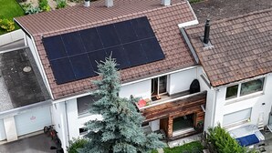 Una casa con un panel solar en el techo, una terraza de madera y un gran árbol de hoja perenne.