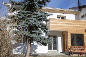 A modern house with a wooden exterior, a large evergreen tree in the foreground, and a bench outside. - CHasa Avant Muglins: Garden flat with a view (Scuol)
