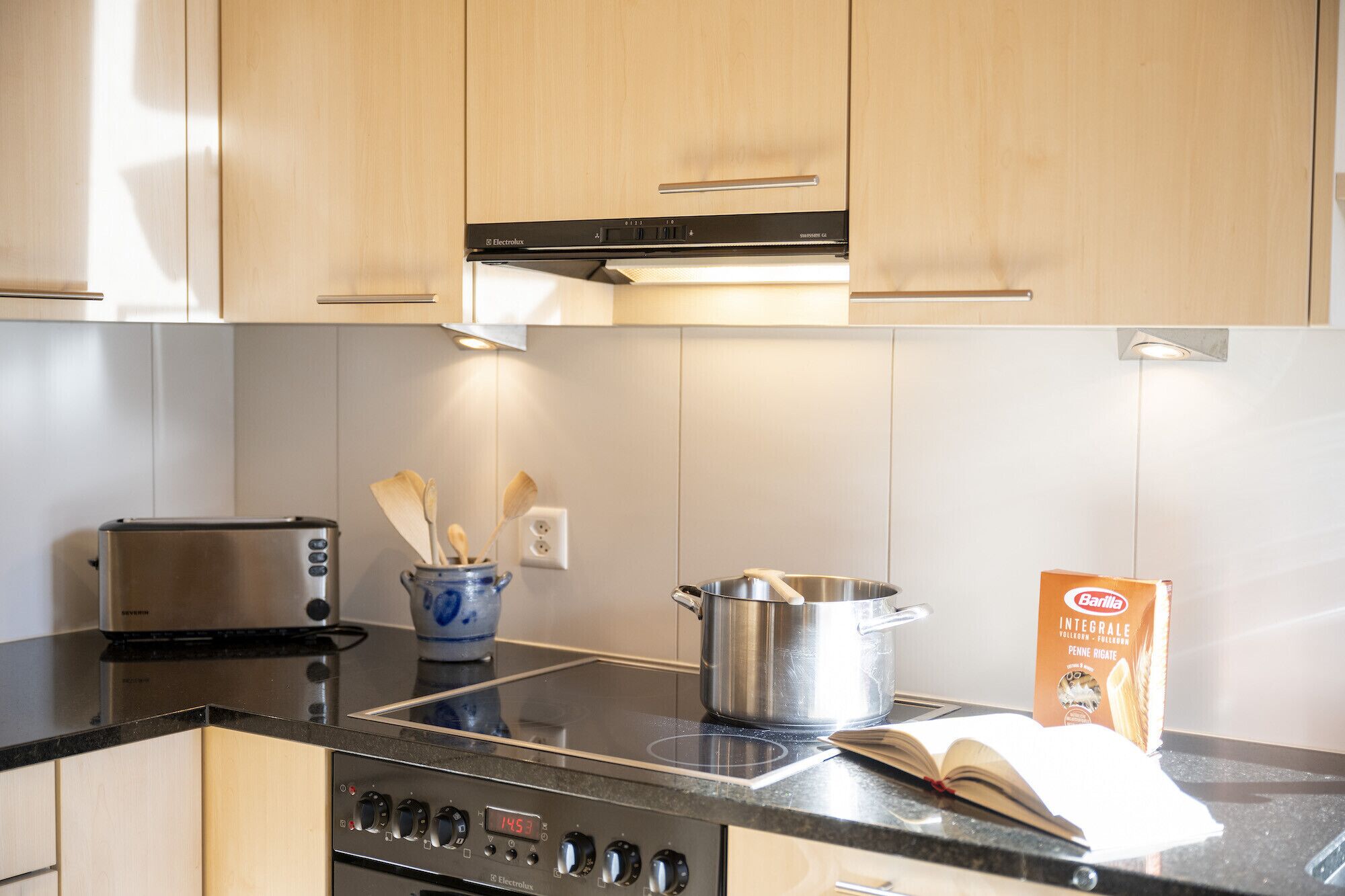 Una cocina moderna con armarios de madera, estufa de acero inoxidable, un tostador, una olla, un libro y un saco de harina.