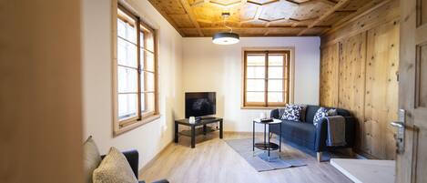Un salón acogedor con piso de madera, un televisor sobre un mueble bajo, un sofá con cojines y ventanas amplias que dejan entrar luz natural.